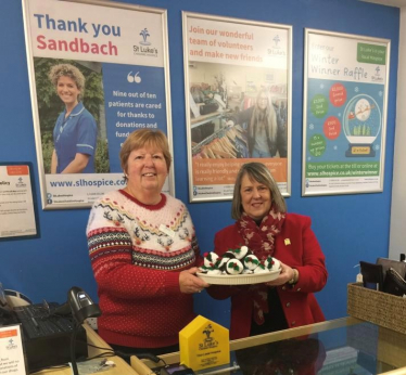 Two women holding a plate of knitted Christmas puddings behind the counter of a St Luke's Hospice shop